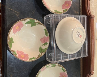 May include: Three off-white ceramic bowls featuring a hand-painted pink flower and green leaf pattern along the interior rim. One bowl is inverted, displaying the Franciscan Desert Rose brand stamp on the base, resting on a clear acrylic stand.