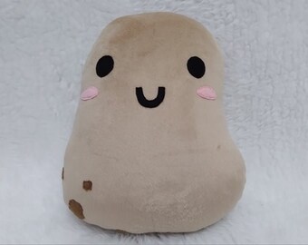 May include: A plush brown potato toy with a smiling face, black eyes, and pink cheeks. The potato has a brown spot on its side.