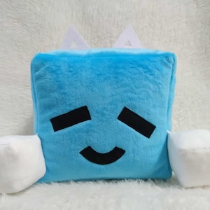 May include: A light blue, cube-shaped plush pillow with black eyes and a curved mouth. Two white, cube-shaped appendages extend from the sides. Two white, triangular shapes are on top of the cube.