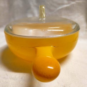 May include: A vintage, yellow glass powder jar with a clear glass lid and a small, clear handle. The jar has a rounded shape with a bulbous base and a small, rounded handle. The jar is sitting on a white surface.