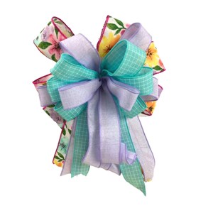 May include: A large, decorative bow made with three different ribbons. The bow is made with a light purple ribbon, a turquoise ribbon with a checkered pattern, and a floral print ribbon with pink, yellow, and green flowers.
