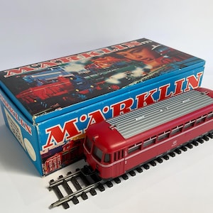 May include: A red and silver toy train engine with a grey roof and black wheels. The engine is on a track and in front of a blue box with the word "MARKLIN" printed on it.