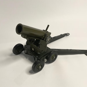 Puede incluir: Un cañón de juguete verde con ruedas negras y un marco de metal. El cañón está sobre un fondo blanco.