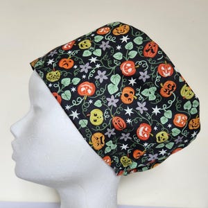 Może przedstawiać: Czarny czepek chirurgiczny z motywem Halloween. Wzór zawiera pomarańczowe i żółte dynie, zielone liście, białe gwiazdy i fioletowe kwiaty. Czepek jest wykonany z tkaniny i jest przeznaczony do noszenia na głowie.