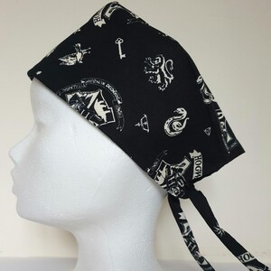 May include: Black scrub hat with white Hogwarts house crests and symbols. The hat has two long ties.