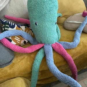 May include: A large, handmade crochet octopus plush toy. The octopus features a teal head and eight long tentacles in shades of pink, blue, and green. The toy has small, round blue eyes and is resting on a yellow couch.