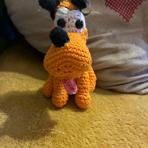 Peut inclure: Un porte-clés fait à la main en forme de chien crocheté orange avec des oreilles et un nez noirs, un visage blanc et une langue rose. Le chien a un porte-clés en métal attaché à sa tête. Le porte-clés est sur une surface jaune.