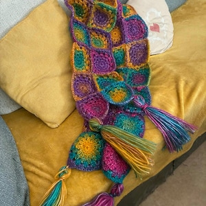 May include: A colorful, handmade crochet scarf with a unique, geometric pattern. The scarf features a mix of vibrant colors including purple, teal, yellow, and pink. Long tassels in matching colors hang from the ends, adding a playful touch. The scarf is displayed on a yellow couch.