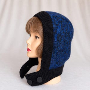 Könnte beinhalten: Eine Strickkapuze mit einem tiefblauen und schwarzen melierten Muster am Oberkopf, eingefasst von einem einfarbigen, schwarzen Rippstrickrand. Die Kapuze verfügt über verlängerte Ohrenklappen, die mit zwei großen, runden, mattschwarzen Knöpfen unter dem Kinn befestigt werden.