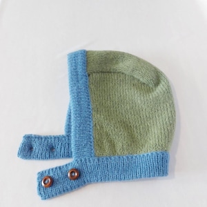 May include: A knitted baby bonnet in shades of green and blue. The main body is olive green, framed with a light blue trim. The bonnet features two blue straps with wooden buttons for closure. The item is displayed on a white background.