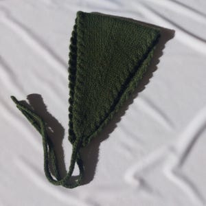 May include: A dark green knitted head covering, triangular in shape with scalloped edges. It features two long knitted ties extending from the bottom corners. The item is displayed on a white surface.