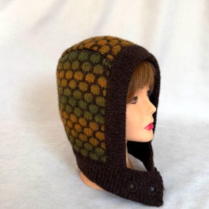 May include: A knitted balaclava featuring a dark brown solid border and a textured honeycomb pattern in shades of olive green and mustard yellow. The headgear covers the top and sides of the head, extending down to the chin with two small buttons.