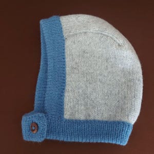 May include: A knitted baby bonnet in gray and blue. The bonnet features a gray main body with a blue border and a button closure. The knitted texture is visible throughout the design, creating a cozy and handmade appearance.