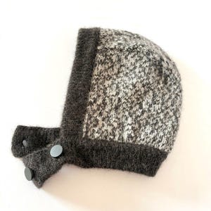 May include: A grey and white knitted baby bonnet with a dark grey trim and two grey buttons. The bonnet is made of a soft, textured yarn and has a rounded shape. The bonnet is designed to keep a baby's head warm.