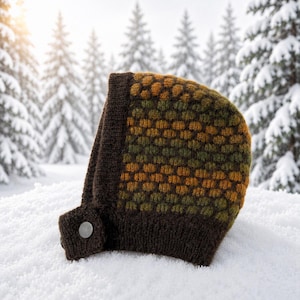 Könnte beinhalten: Eine gestrickte Haube mit einem strukturierten Wabenmuster in Senfgelb, Olivgrün und Dunkelbraun. Die Kopfbedeckung hat einen einfarbigen, dunkelbraunen Ripprand und eine kleine seitliche Lasche, die mit einem silbernen Metallknopf befestigt ist.