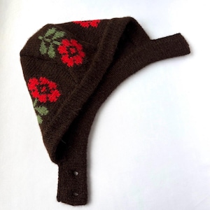 May include: A dark brown knitted bonnet featuring a repeating floral pattern in bright red and sage green. The headwear includes a wide brim, a structured crown, and a long chin strap with two circular buttonholes for secure fastening under the chin.