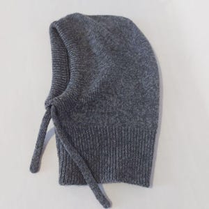 May include: A gray knit balaclava with a ribbed bottom and long ties. The balaclava is made of a soft, warm material, perfect for cold weather. The hood covers the head and neck.