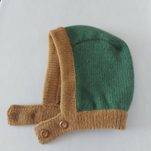 May include: A knitted baby bonnet in green and tan. The bonnet has a green top and tan trim and straps. The straps fasten with two wooden buttons. The bonnet is made of a soft, textured yarn.