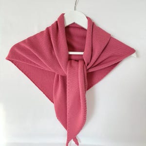 Puede incluir: Un chal de punto color rosa empolvado exhibido en una percha blanca. El chal es triangular, con una sección larga y rectangular colgando. El tejido de punto tiene una textura sutil y los bordes están cuidadosamente acabados. Un accesorio suave y cálido.