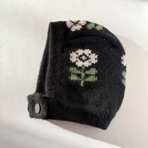 Puede incluir: Un gorro negro de punto con un diseño floral. El gorro presenta flores blancas y verdes. Tiene un cierre de botón en el puño. El gorro está hecho de un material suave y texturizado.