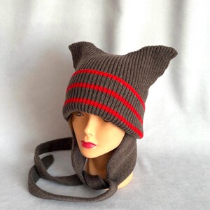 May include: A dark gray knit beanie featuring two pointed cat-ear shapes at the top. The hat includes two horizontal bright red stripes around the cuff area and two long, dangling knit ties extending from the sides to secure under the chin.