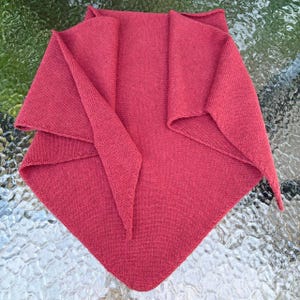 May include: A triangular knit shawl in a solid deep red color. The garment features a fine, consistent stockinette stitch texture throughout, with folded edges that create a layered appearance at the top corners and a pointed bottom hem.