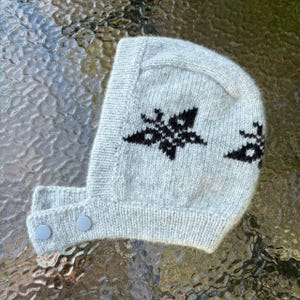 Könnte beinhalten: Eine hellgraue Strickhaube mit einem schwarzen Sternenmuster. Die Kopfbedeckung hat eine strukturierte Kapuzenform mit einem Kinnriemen, der mit zwei runden, hellgrauen Knöpfen befestigt wird. Die Strickstruktur ist deutlich erkennbar.