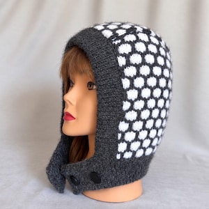 May include: A knit balaclava featuring a dark charcoal gray solid border and a white and dark charcoal gray honeycomb pattern on the crown. The headgear includes two black circular buttons at the chin area to secure the fit around the face.