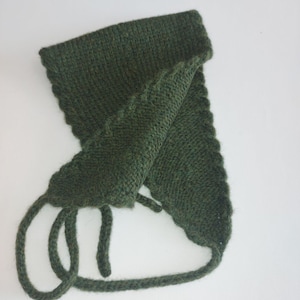 Può includere: Un cappello lavorato a maglia verde scuro con paraorecchie e lacci. Il cappello è realizzato con un filato strutturato e ha un bordo smerlato. I lacci sono lunghi e ad anello. Il cappello è probabilmente fatto a mano e adatto al clima freddo.