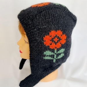 Peut inclure: Un bonnet noir en tricot avec un motif floral orange et vert. Le bonnet a des cache-oreilles et une mentonnière. La fleur est sur le côté du bonnet. Le bonnet est sur une tête de mannequin.