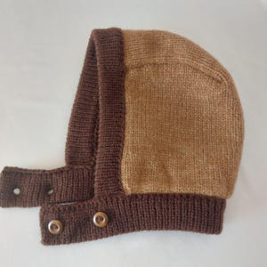 May include: A knitted baby bonnet in shades of brown. The bonnet features a light brown center panel framed by a darker brown border and straps. The straps have button closures. The bonnet is made of soft yarn.