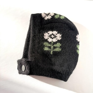 Puede incluir: Un gorro negro tejido con un diseño floral. El gorro presenta una flor blanca con hojas verdes. Tiene un cierre de botón y está hecho de un material suave y texturizado. El gorro es un accesorio de invierno.
