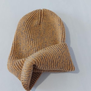 Könnte beinhalten: Senfgelbe Strickmütze mit umgeschlagenem Saum. Die Mütze besteht aus einem Rippenstrickstoff mit grauen Sprenkeln. Ein warmes, bequemes Accessoire für kaltes Wetter.