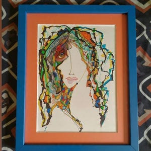 May include: An abstract portrait drawing in a blue frame with an orange mat. The artwork features a woman's face with colorful, flowing hair and details in shades of green, orange, yellow, and blue. The drawing is on white paper.