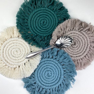 May include: Four round crocheted coasters with fringe, in shades of green, beige, white, and blue. A silver serving spoon is placed on top of the coasters.