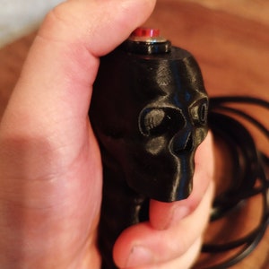 Puede incluir: Botón negro con forma de calavera con un botón rojo en la parte superior. La calavera tiene rasgos faciales detallados y se sostiene en una mano. Un cable negro se extiende desde la base de la calavera.