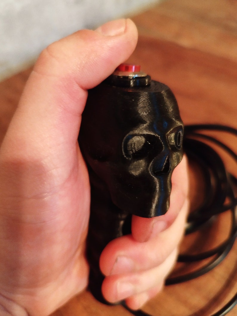 May include: Black skull-shaped button with a red button top. The skull has detailed facial features and is held in a hand. A black cord extends from the base of the skull.