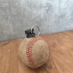Könnte beinhalten: Ein abgenutzter Baseball mit roten Nähten und einem schwarzen Aufsatz mit einem silbernen Ring. Er liegt auf einer Holzoberfläche. Der Hintergrund ist hellgrau.