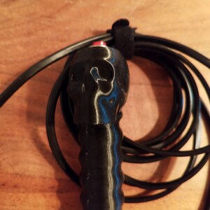 May include: A black skipping rope featuring a skull-shaped handle. The handle has a red button and a textured grip. The rope is coiled and secured with a black velcro strap. The background is a wooden surface.