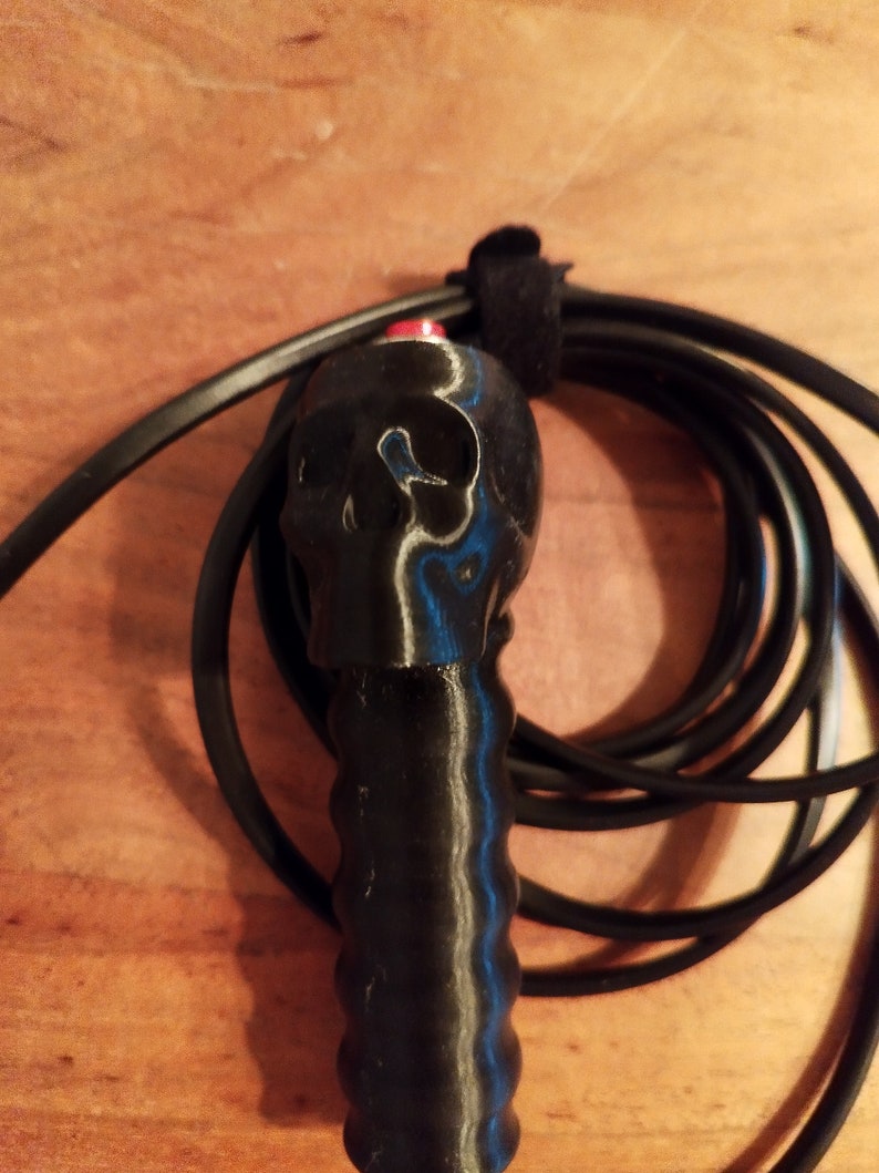 May include: A black skipping rope featuring a skull-shaped handle. The handle has a red button and a textured grip. The rope is coiled and secured with a black velcro strap. The background is a wooden surface.