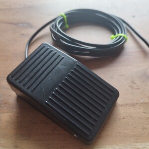 May include: A black foot pedal switch with a long black cable and a green tie wrap. The pedal has a textured surface.