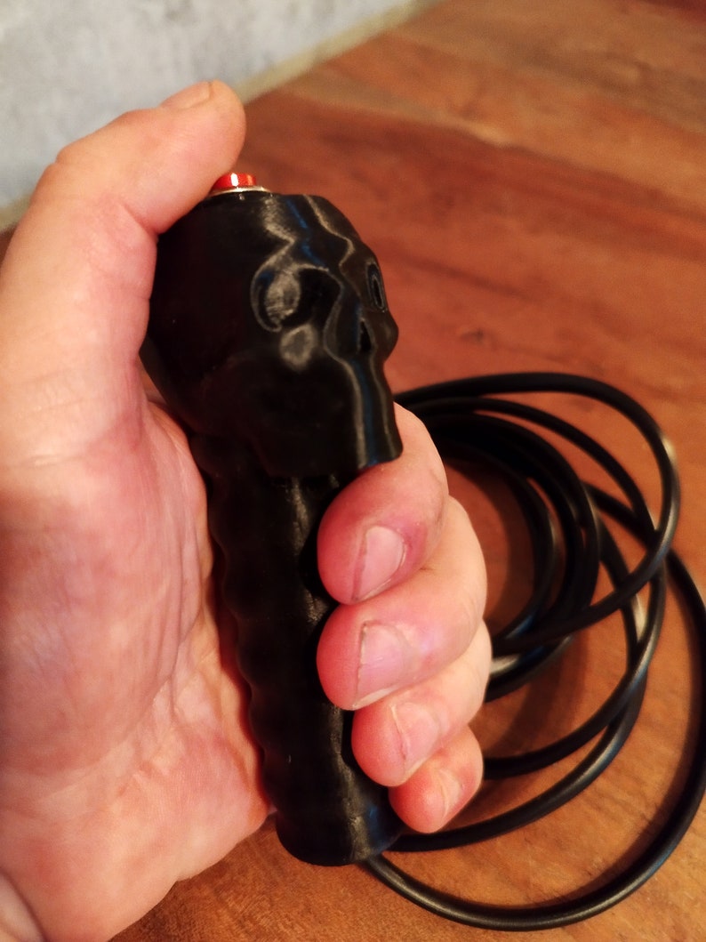May include: Black skull-shaped handle with a red button. The handle is connected to a black cable. The product is held in a hand.