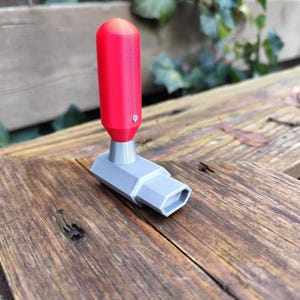 May include: A 3D-printed tool with a red cylindrical handle and a gray rectangular base. The tool is resting on a weathered wooden surface, with green foliage in the background. The handle is approximately 2 inches long.