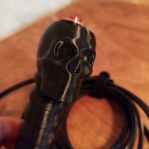May include: A black skull-shaped handle with a red button. The handle is connected to a black cable. The skull features eye sockets and a nose cavity. The handle has a textured grip.