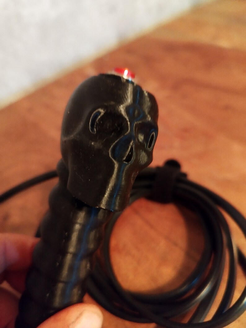 May include: A black skull-shaped handle with a red button. The handle is connected to a black cable. The skull features eye sockets and a nose cavity. The handle has a textured grip.