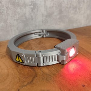 Könnte beinhalten: Ein graues, kreisförmiges Gerät mit einer rechteckigen Komponente mit hellem rotem Licht. Das Gerät hat ein Scharnierdesign und ein gelbes Gefahrensymbol. Das Objekt befindet sich auf einer Holzoberfläche.