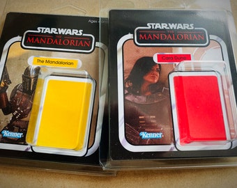 May include: Two Star Wars The Mandalorian action figure packaging boxes. The left box features The Mandalorian in a yellow background. The right box features Cara Dune in a red background. Both boxes have the Kenner logo.