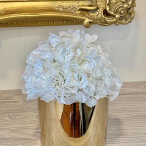 May include: A gold-colored, cylindrical vase with a reflective surface, filled with a cluster of white flowers. The vase sits on a light-colored wooden surface, with a gold-framed object in the background.