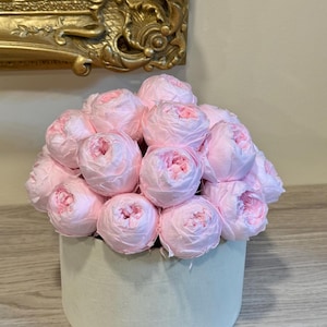 May include: A collection of light pink roses arranged in a round, light green box. The roses are tightly packed, with delicate petals. The box sits on a wooden surface, with a gold framed picture in the background.