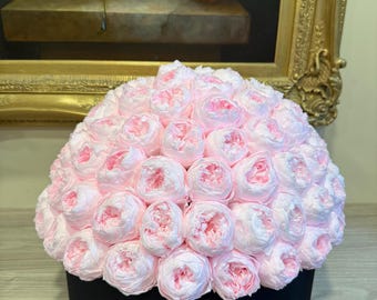 May include: A round black box overflowing with light pink and white paper flowers. The flowers are arranged in a dense, dome-like shape. The background includes a painting in a gold frame.